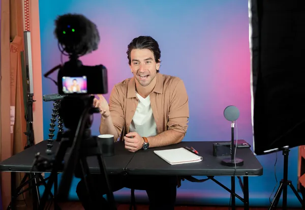 Male content creator recording video at desk with professional camera, ring light, and colorful gradient background studio setup