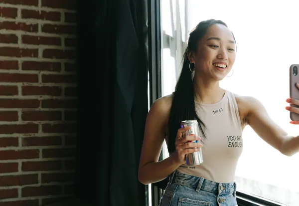 Female influencer holding branded can, taking selfie video content by window with brick wall